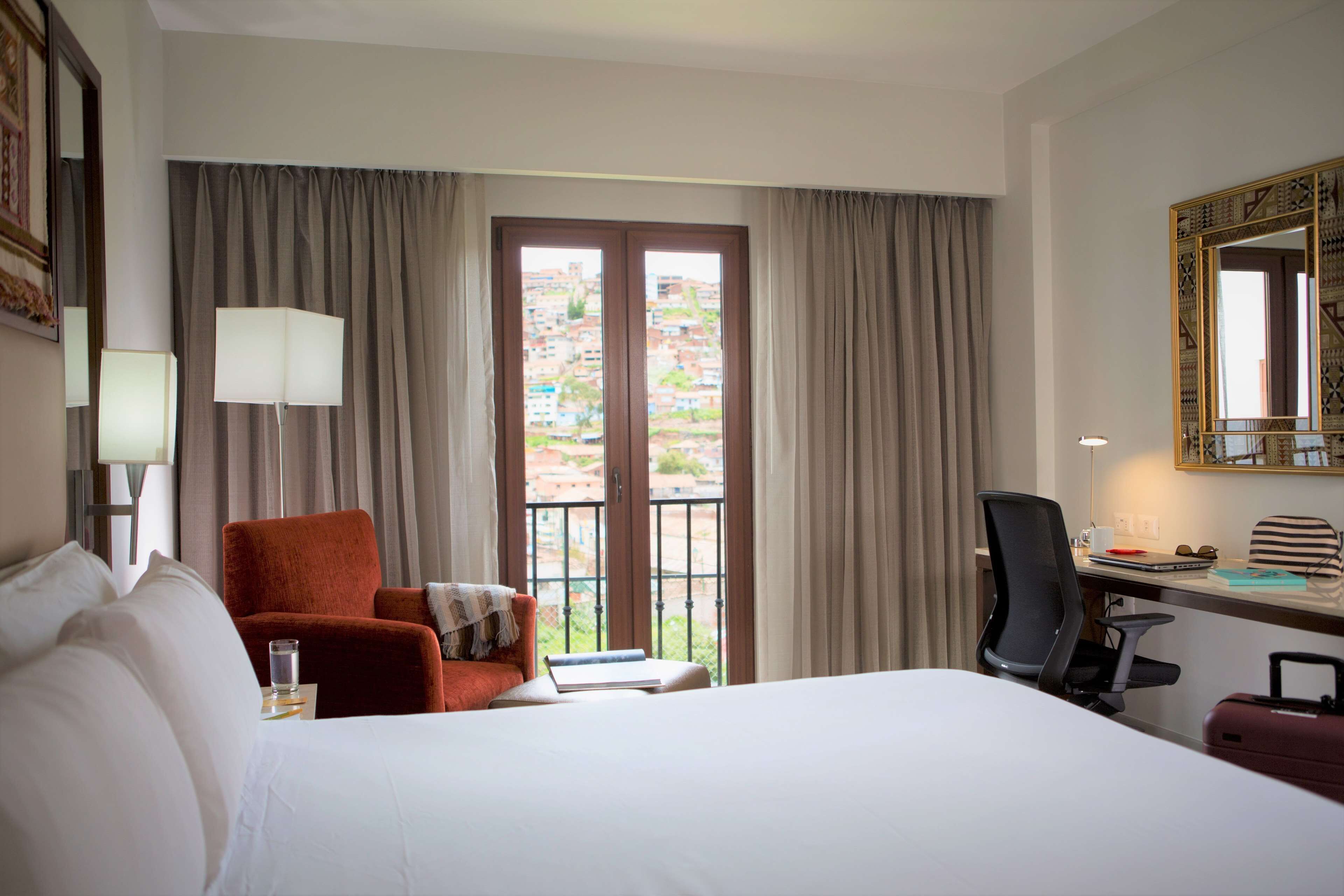 A hotel room with a bed, a desk with a chair, a red armchair, a window with curtains, and a large mirror on the wall.