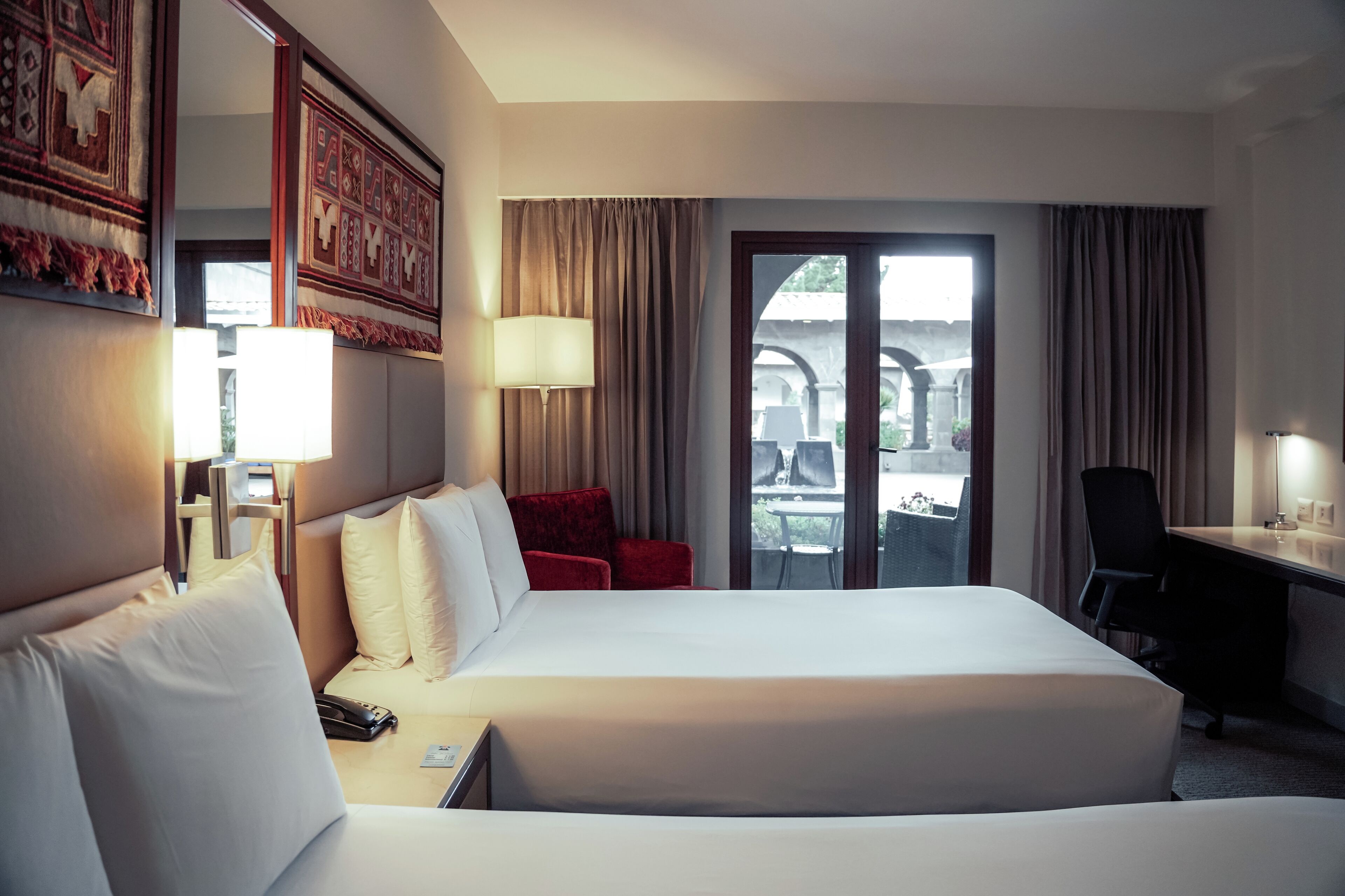 A hotel room with a large bed, a desk, a chair, and a balcony with a view of the outside.
