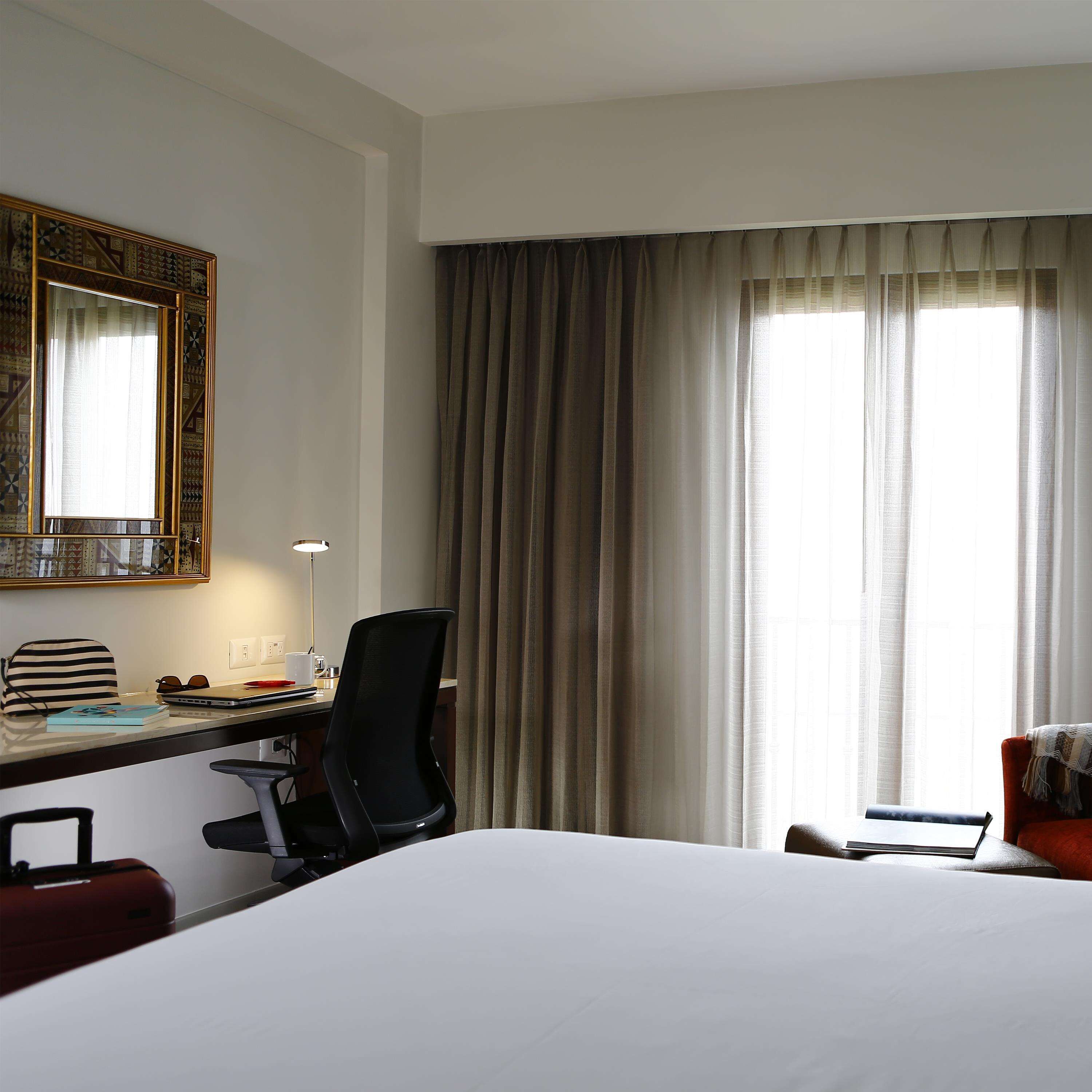 Quarto de hotel com uma cama, uma escrivaninha com um laptop, uma cadeira, uma mala e uma janela grande com cortinas.