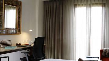Quarto de hotel com uma cama, uma escrivaninha com um laptop, uma cadeira, uma mala e uma janela grande com cortinas.