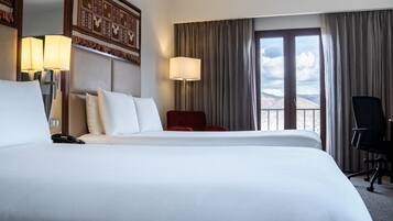 A hotel room with two beds, a red chair, a desk, and a view of mountains through the window.