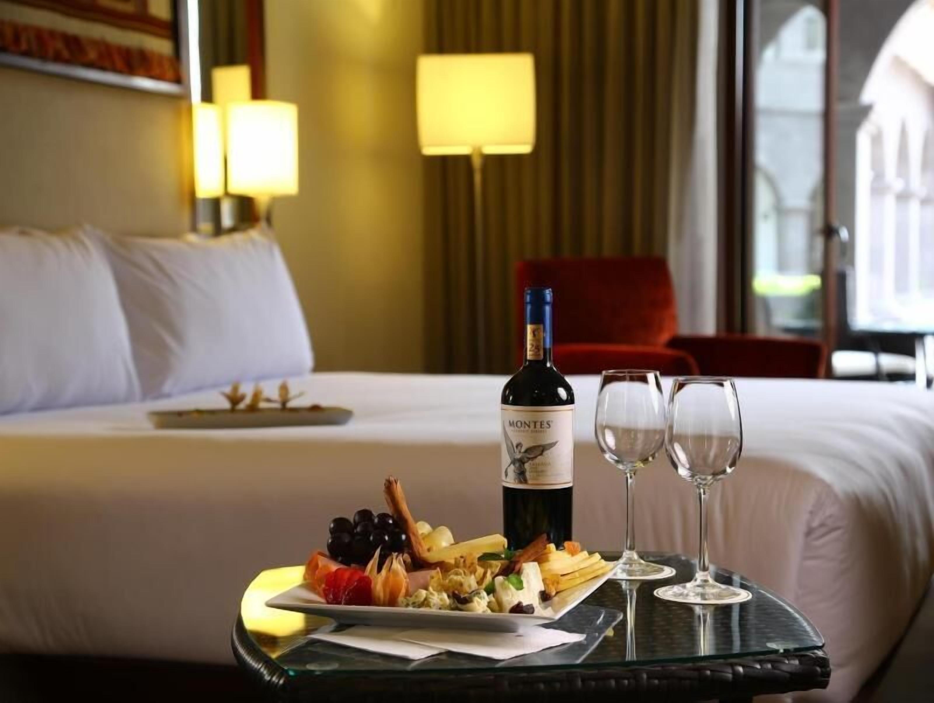 A hotel room with a bed, a tray with food and wine, and two glasses.