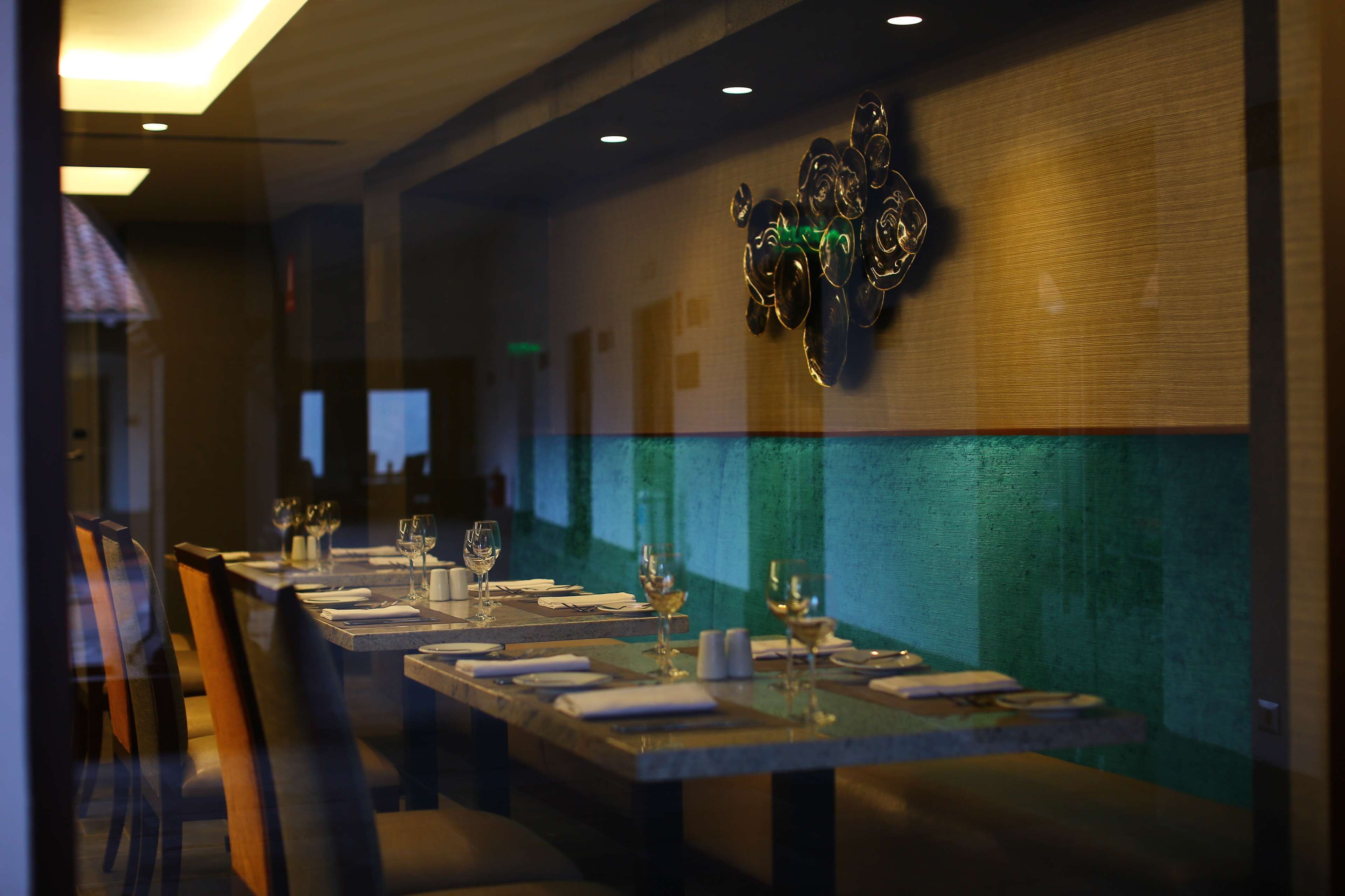 A restaurant with long tables set for dining, a green textured wall, and a decorative metal piece above the tables.