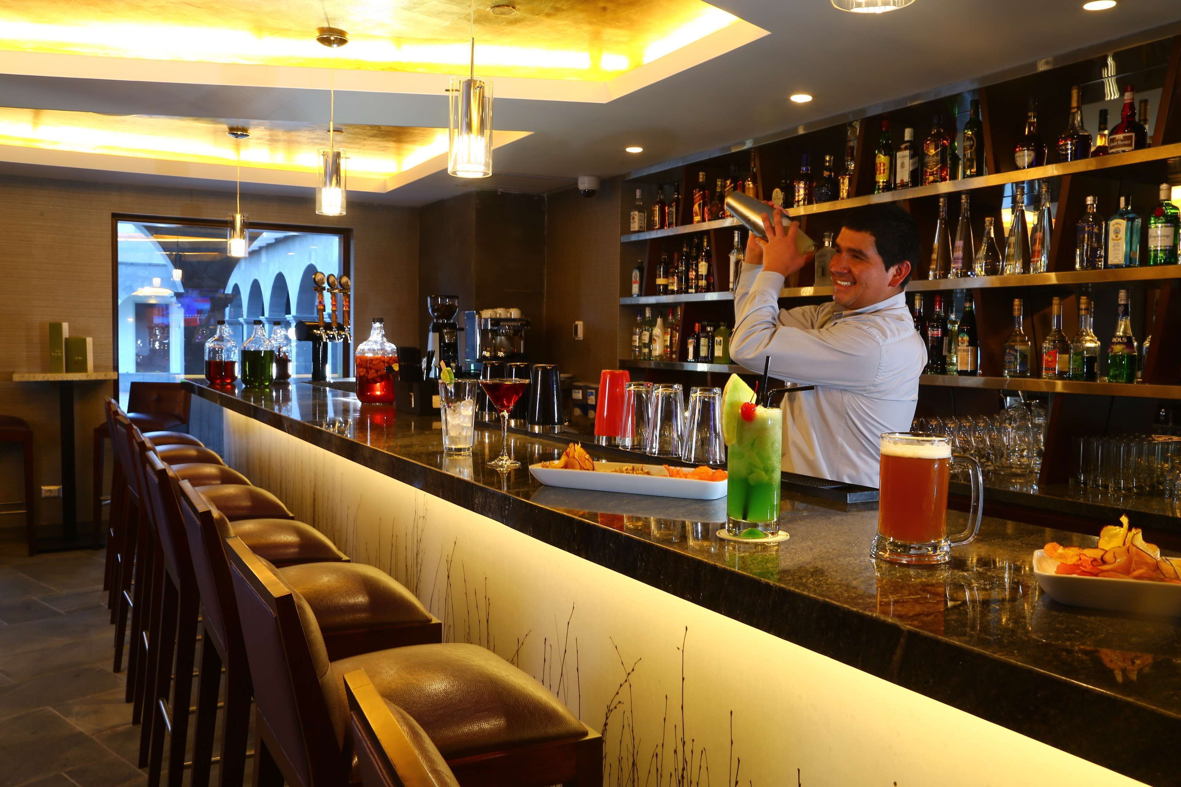 A bar area with a bartender preparing drinks, a variety of bottles on shelves, and a seating area with chairs.