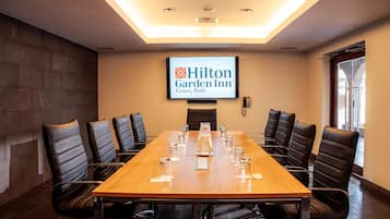Un salón de conferencias con una larga mesa, sillas de cuero negro, una pantalla de proyector mostrando el logotipo de Hilton Garden Inn y un teléfono en la pared.