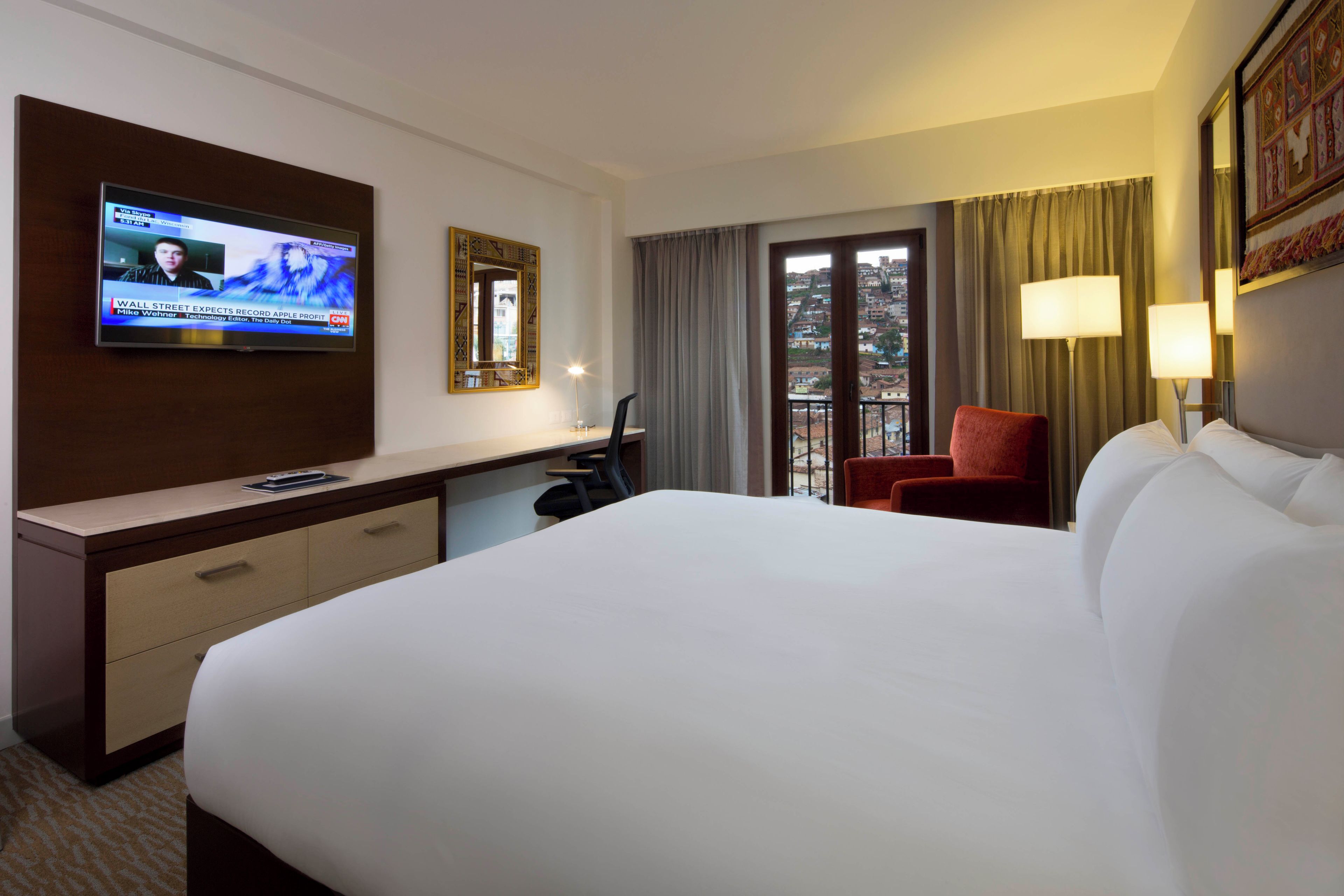 A hotel room with a large bed, a desk with a chair, a TV on the wall, a window with curtains, and a door leading to an outdoor area.