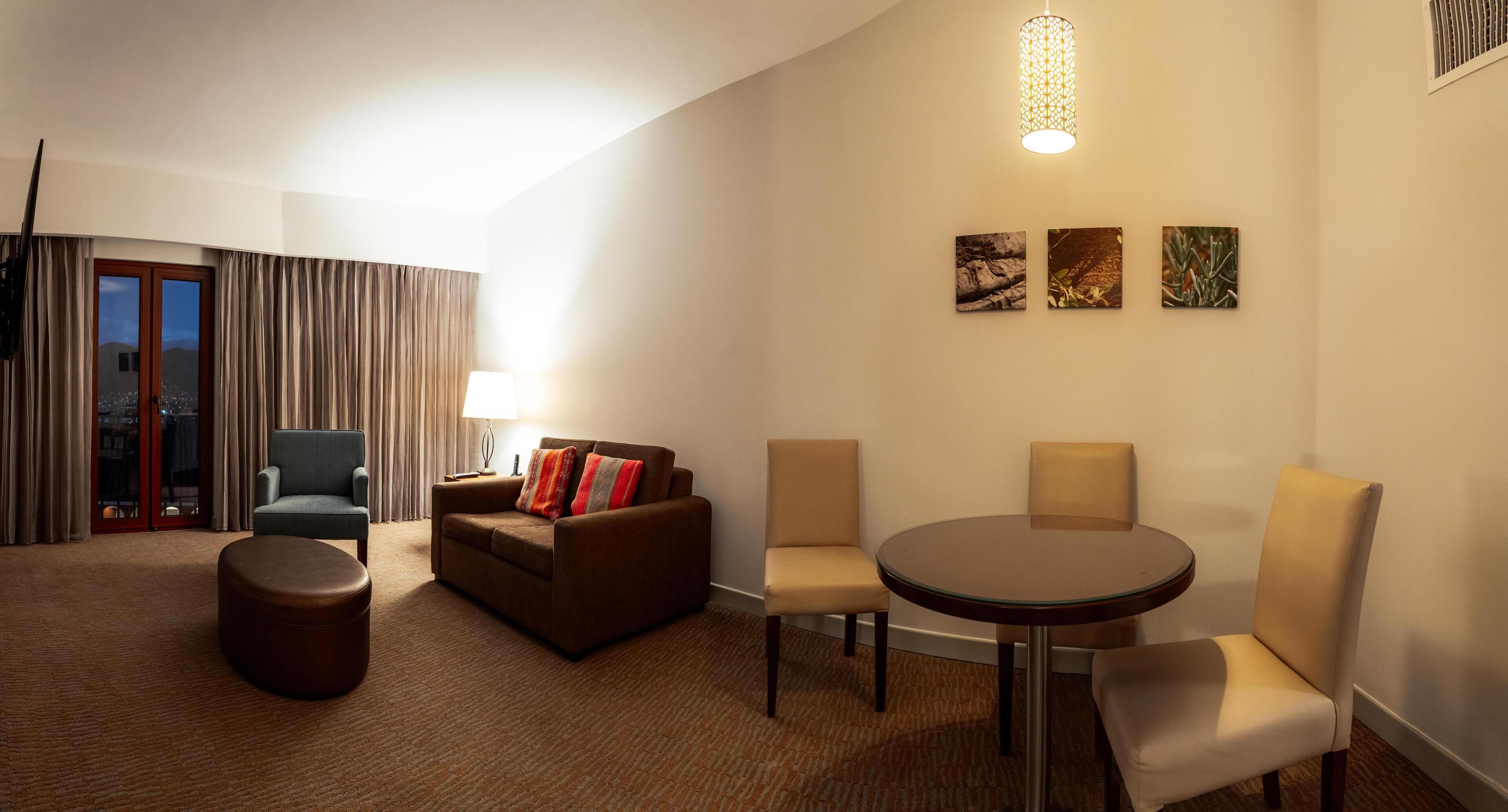 A hotel room with a sofa, two chairs, a round table, a lamp, and three wall-mounted pictures.