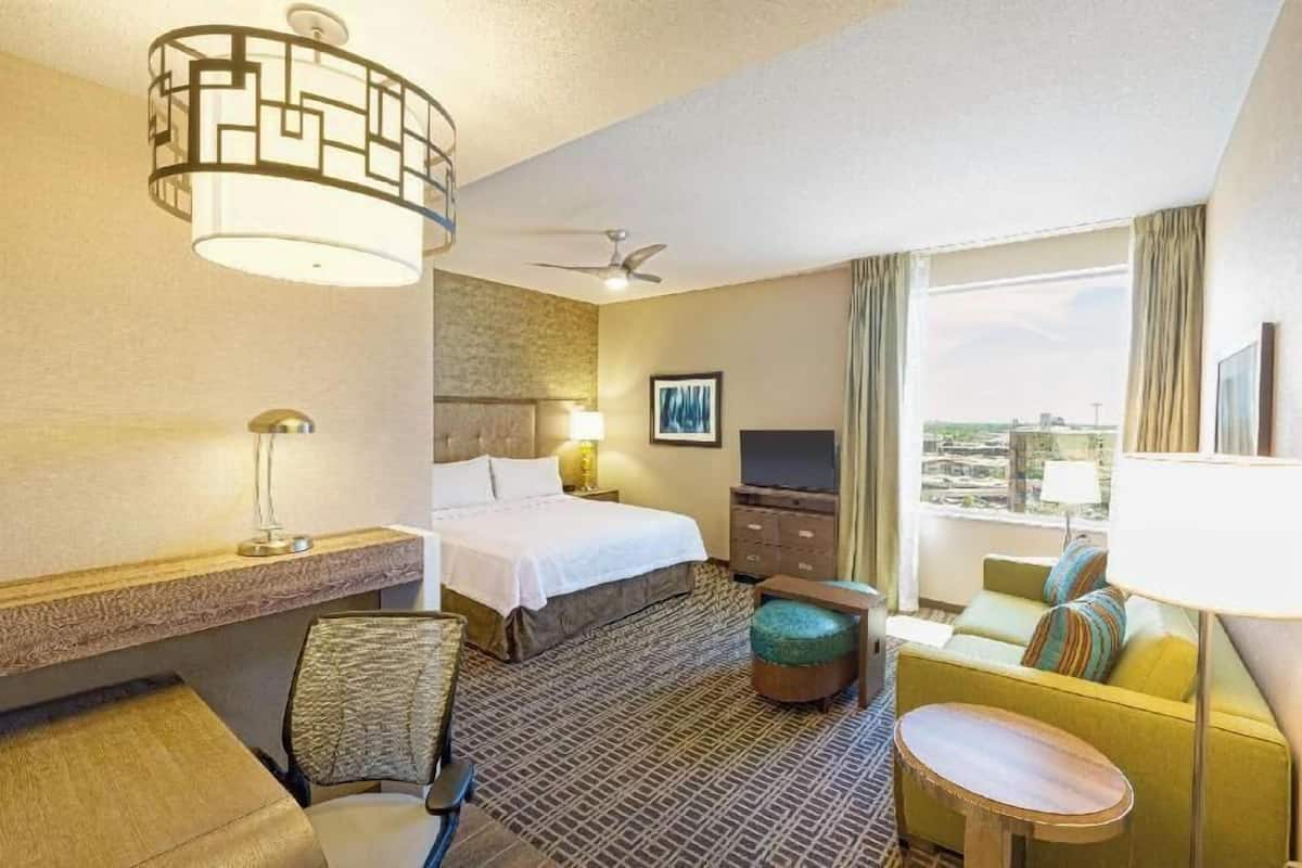 A hotel room with a bed, a desk, a chair, a sofa, a TV, a window with curtains, and a ceiling fan.