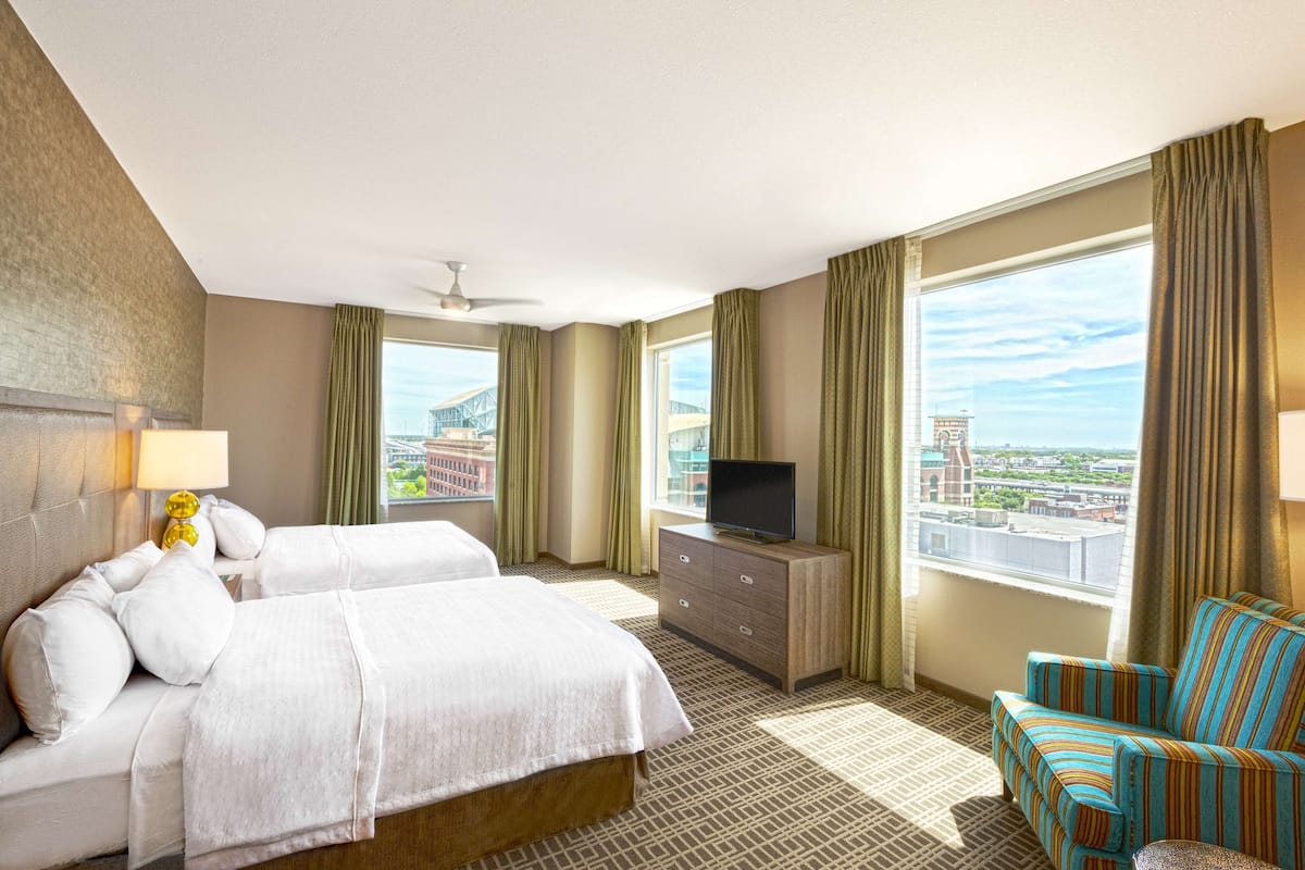 A hotel room with two beds, a TV on a dresser, a chair, and a large window with a city view.