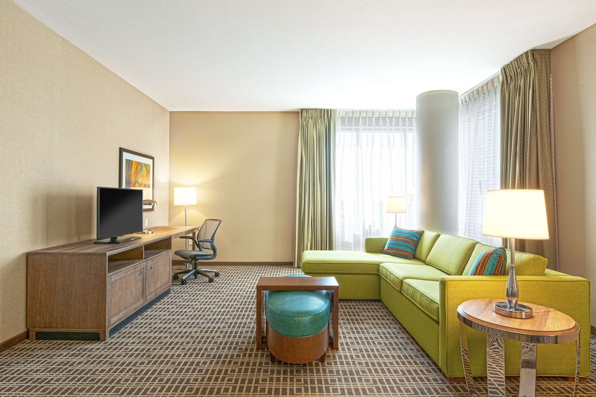 A hotel room with a green sofa, a wooden desk with a flat-screen TV, a small round table with a lamp, and a patterned carpet.