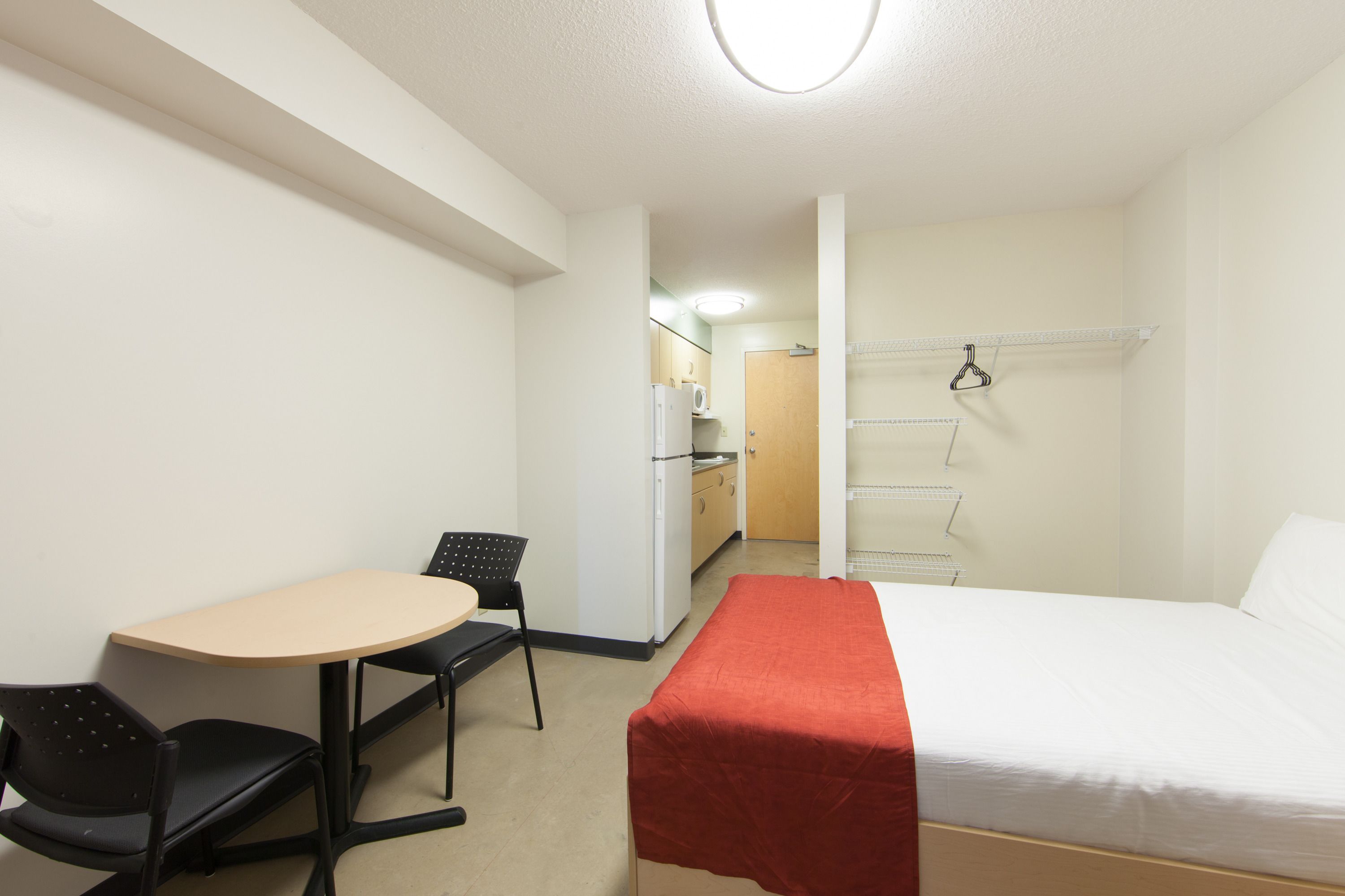 A hotel room with a bed, a desk with a chair, a kitchenette, and a door leading to another room.