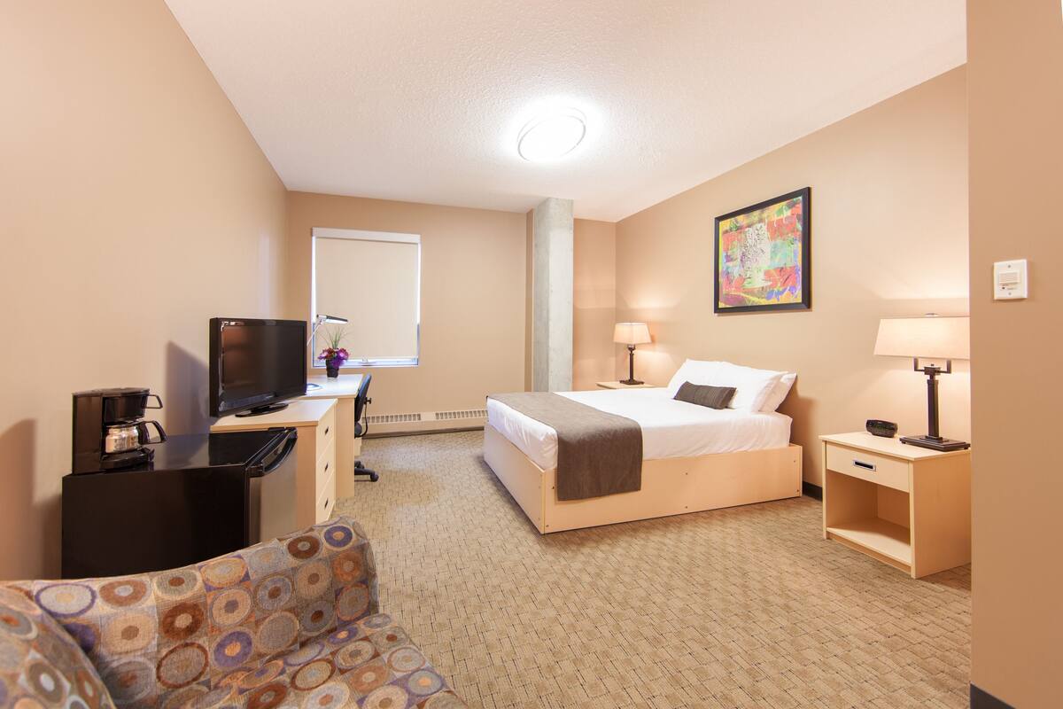 A hotel room with a bed, a desk with a TV, a chair, a lamp, and a painting on the wall.