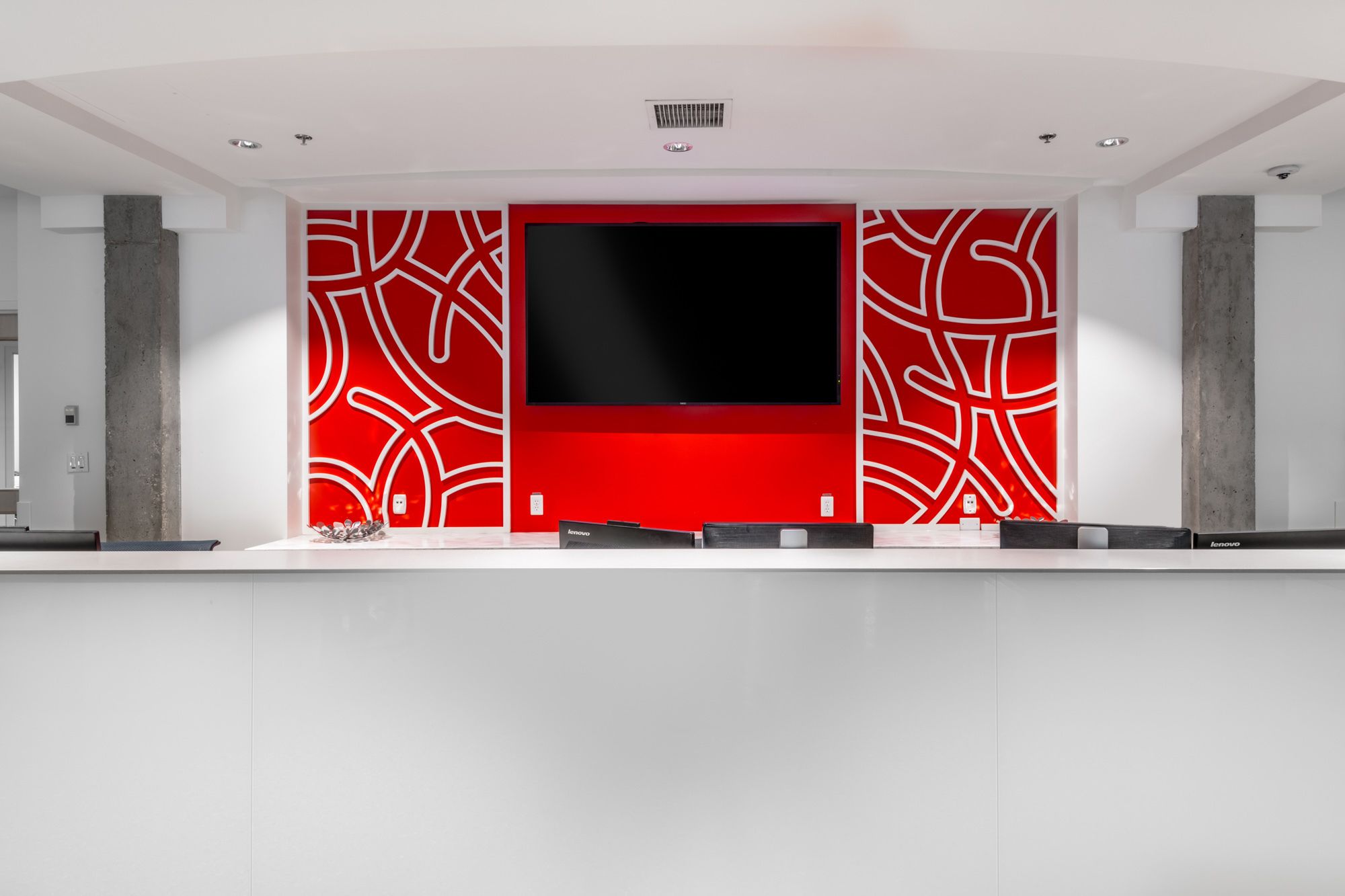 A modern reception desk with a large flat-screen TV, red patterned walls, and a minimalist design.