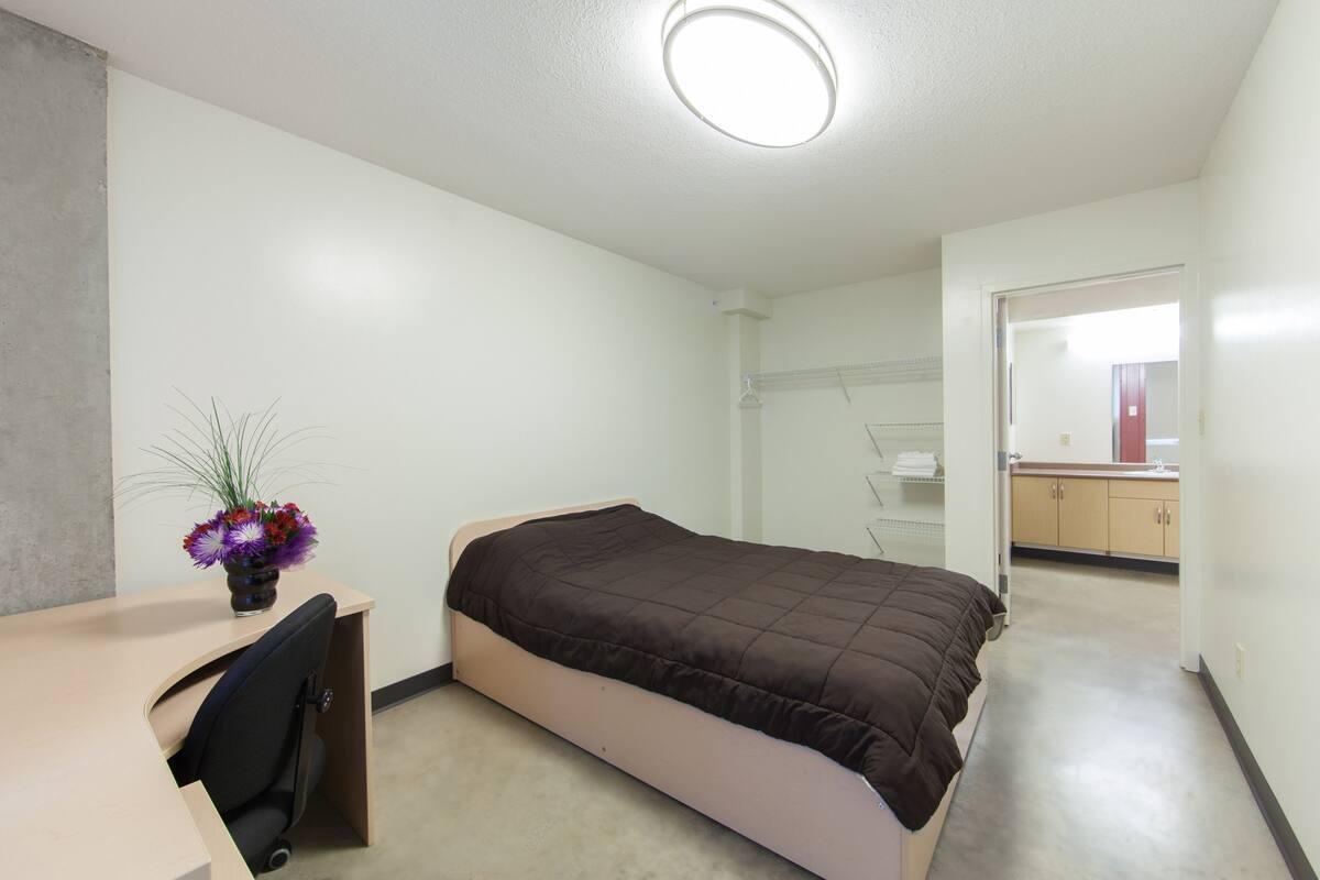 A bedroom with a bed, a desk, and a chair. There is a bathroom visible through an open door.