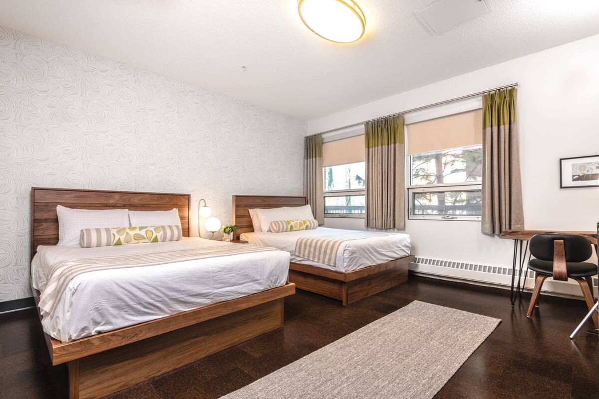 A hotel room with two wooden beds, a small table, a chair, and a large window with curtains.