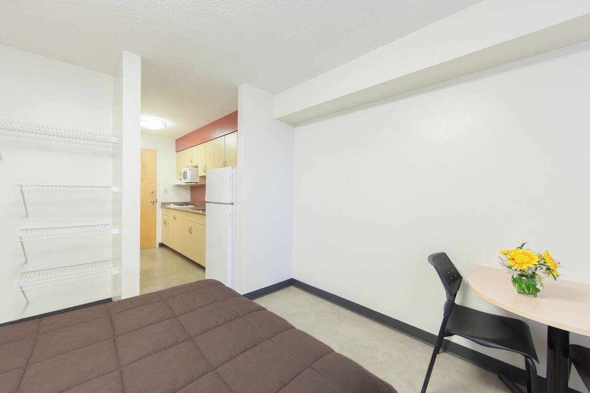 A hotel room with a bed, a dining table with a chair, a kitchen area, and a door leading to another room.