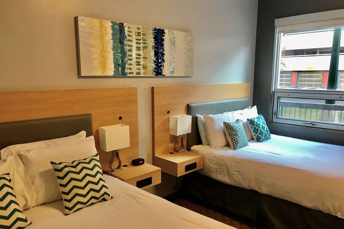 A hotel room with two beds, a nightstand with a lamp, a bedside table with a clock, and a window with a view of buildings.