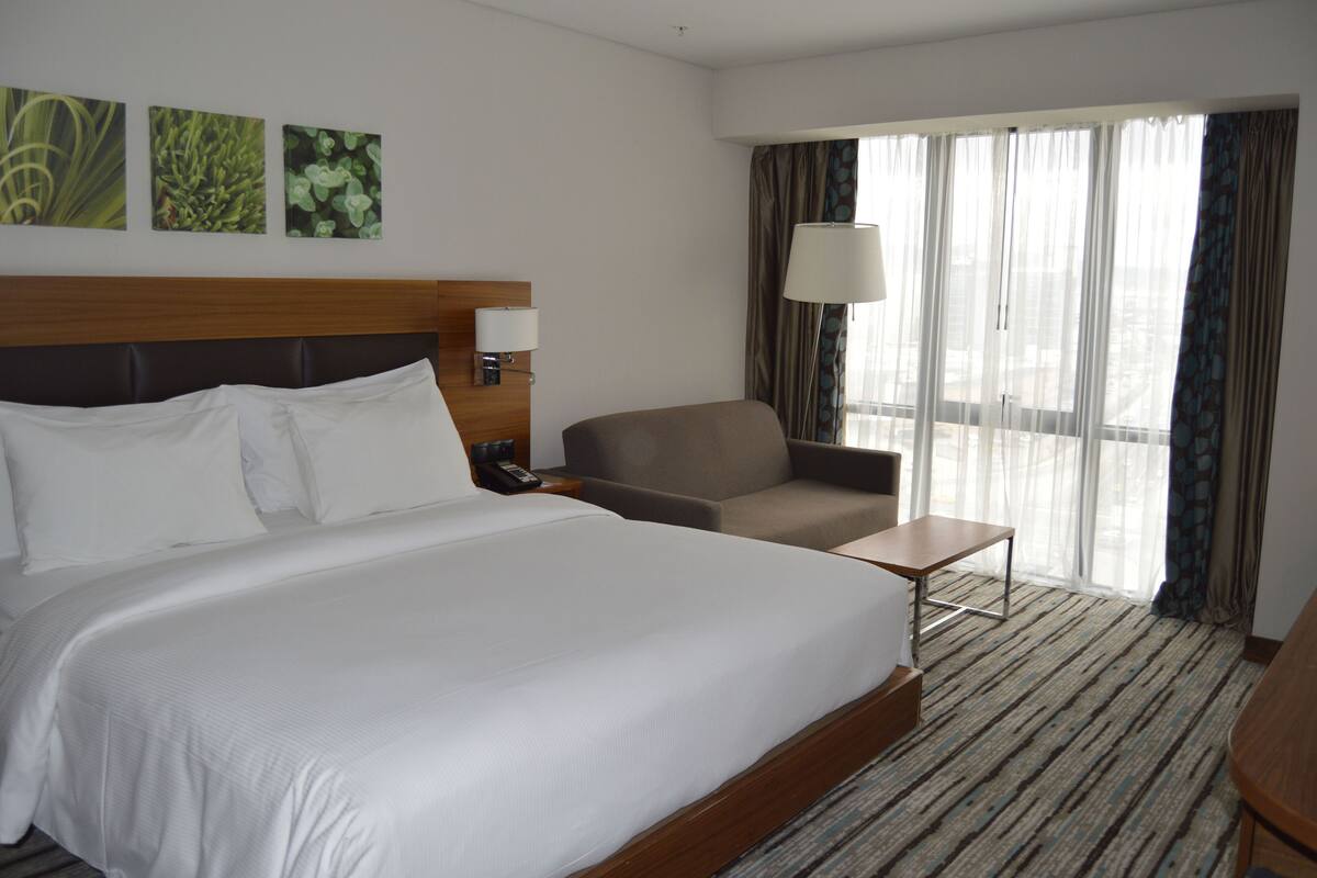 Une chambre d’hôtel avec un grand lit, une chaise, une petite table et une fenêtre avec des rideaux.