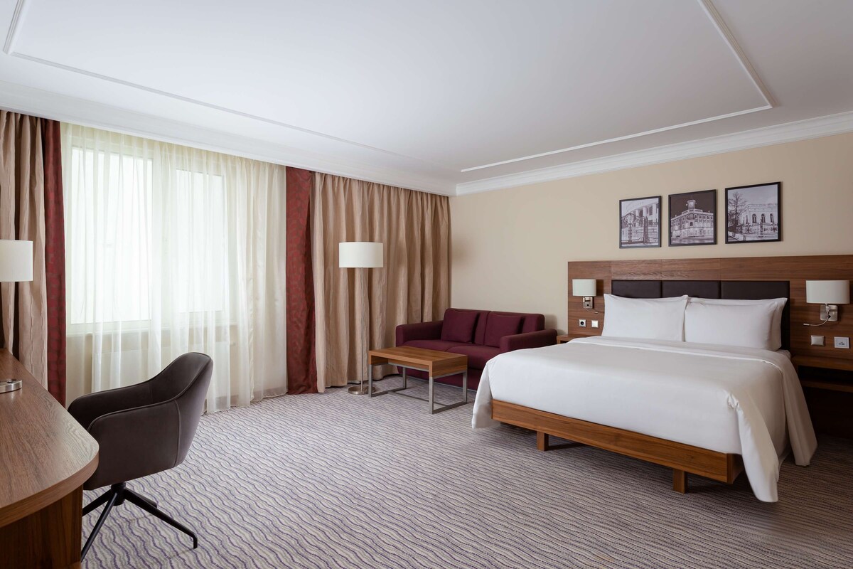 Una habitación de hotel con cama, escritorio, silla, sofá y ventana con cortinas.