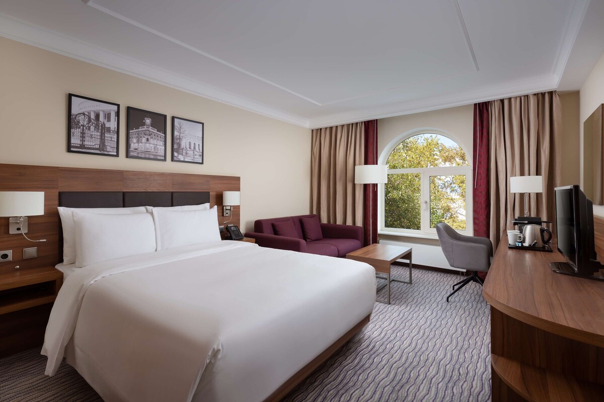 Una habitación de hotel con una cama grande, un escritorio con una silla, un sofá y un televisor. Hay cuadros enmarcados en la pared y una ventana con cortinas.
