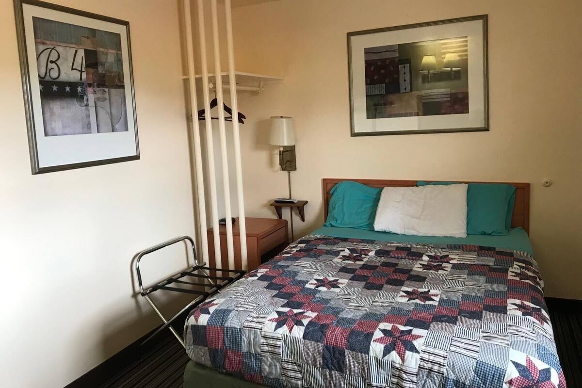 A hotel room with a bed, a chair, a nightstand, a lamp, and two framed pictures on the walls.