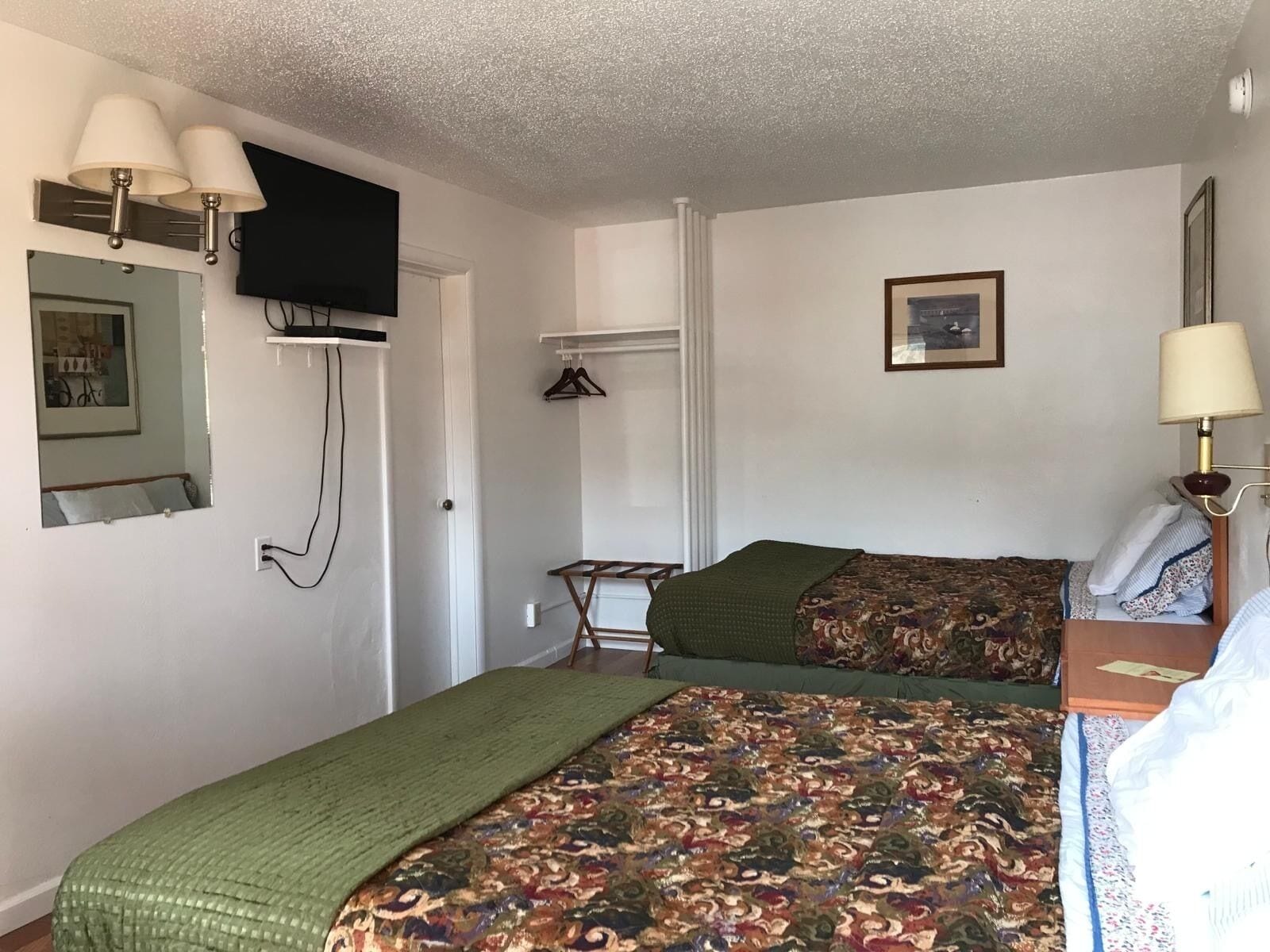 Una habitación con dos camas, un televisor montado en la pared, un perchero y un cuadro enmarcado.