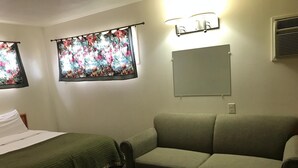 A hotel room with a bed, a sofa, a window with floral curtains, a wall-mounted mirror, and an air conditioning unit. - Blue Mountain Hotel (Dayton)