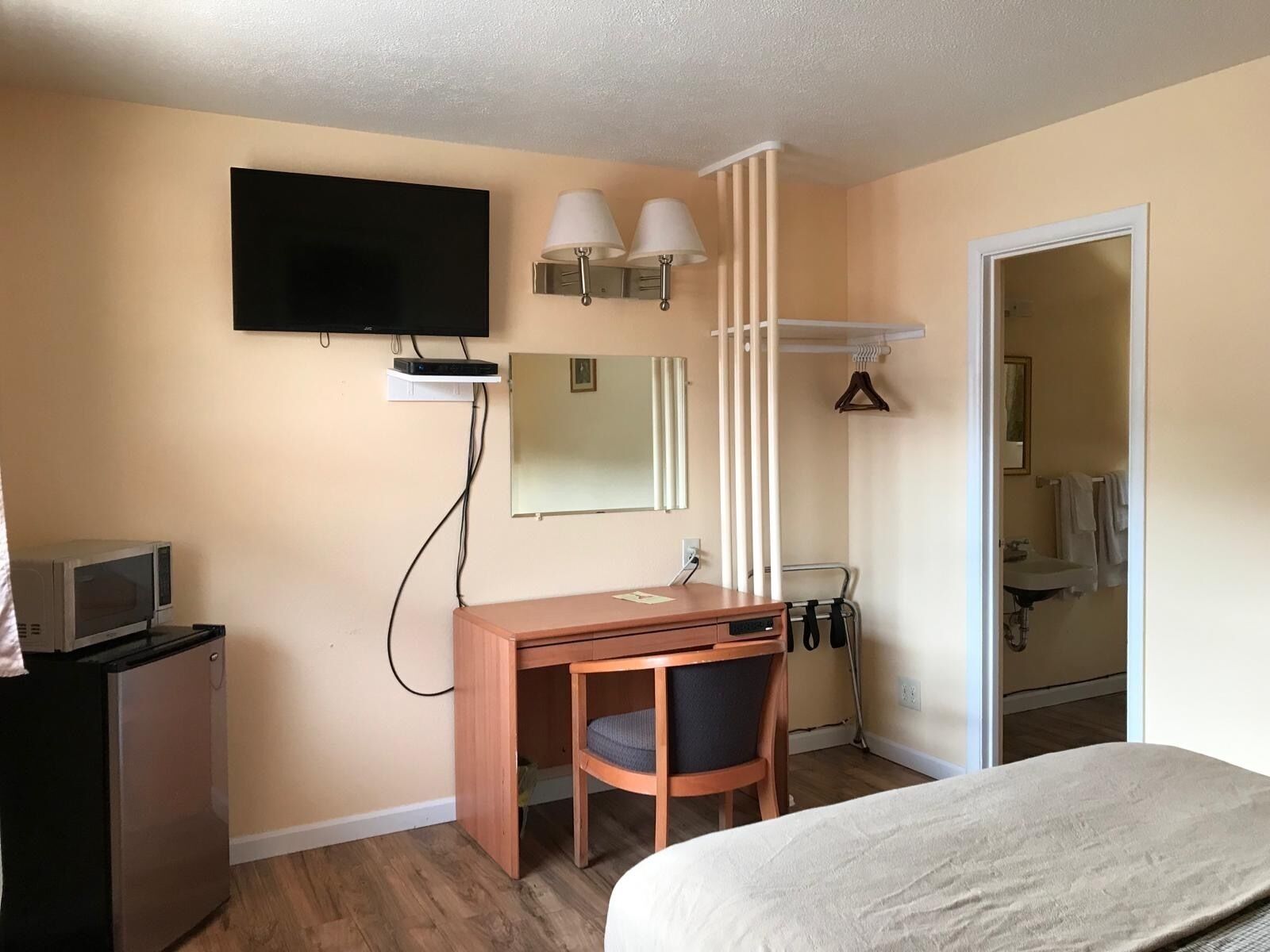 Habitación de hotel con escritorio de madera, una silla, cama, televisor, espejo y baño visible a través de una puerta abierta.
