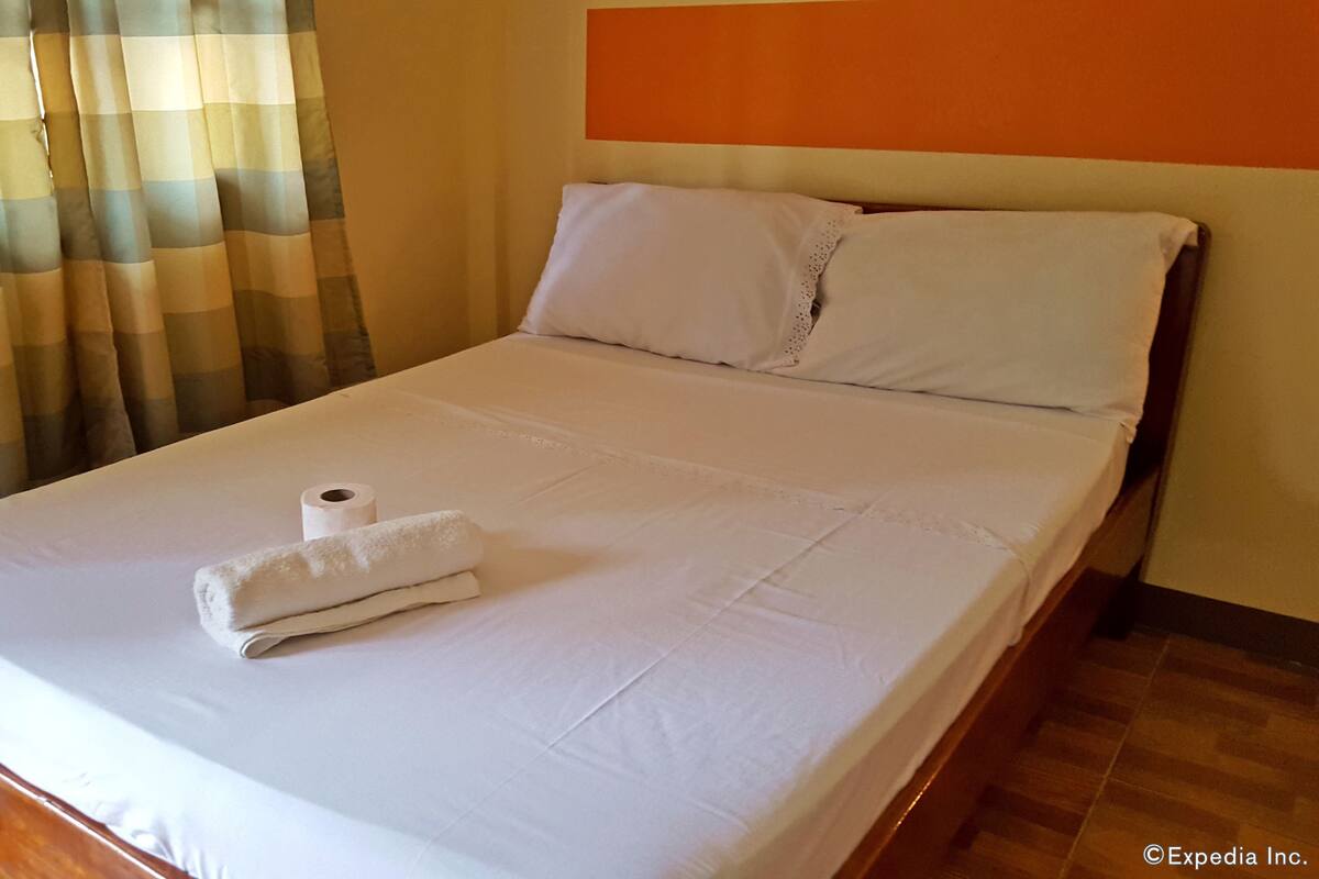 A neatly made bed with white linens, a rolled-up towel, and a roll of toilet paper on the bed.