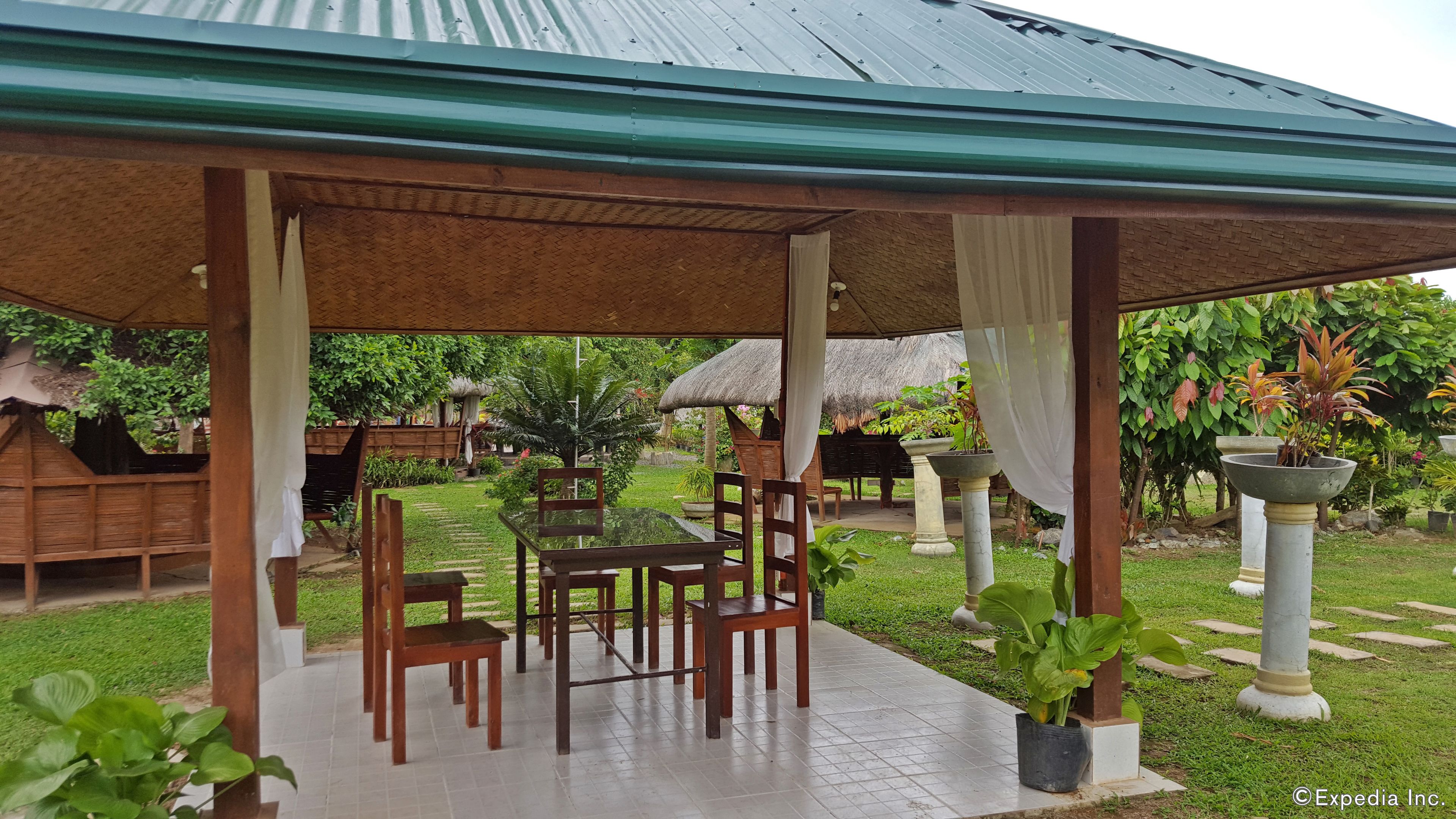 A covered outdoor seating area with wooden tables and chairs, surrounded by lush greenery and a thatched-roof structure.
