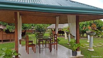 A covered outdoor seating area with wooden tables and chairs, surrounded by lush greenery and a thatched-roof structure.