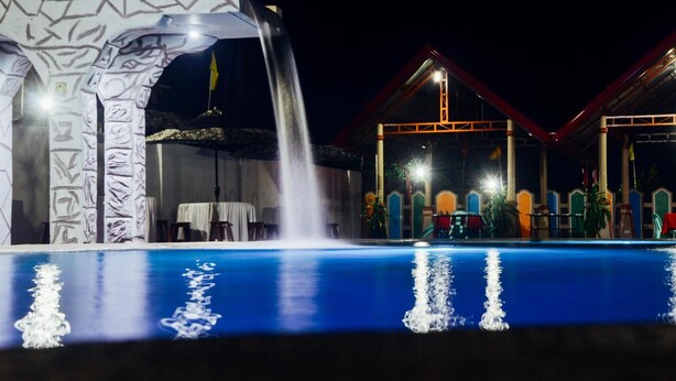 Una piscina iluminada en la noche con una cascada, rodeada por un edificio de arquitectura colorida y un área techada con mesas y sillas.