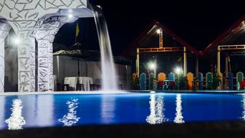 A nighttime swimming pool with a waterfall feature, surrounded by a building with colorful architecture and a covered area with tables and chairs.