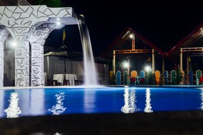 A nighttime swimming pool with a waterfall feature, surrounded by a building with colorful architecture and a covered area with tables and chairs. - Michelle Pension (Puerto Princesa)