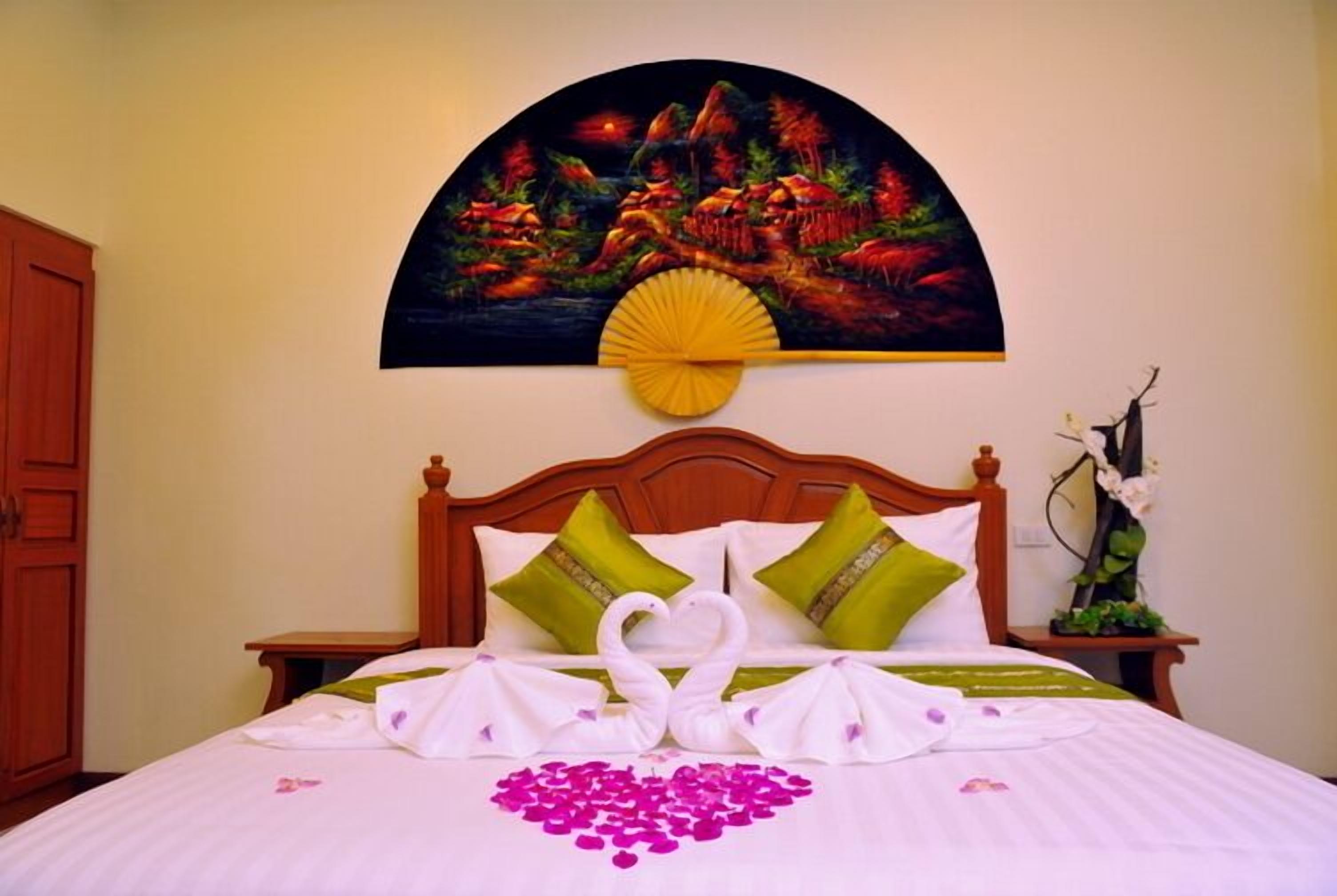 A neatly made bed with a heart-shaped arrangement of rose petals and swan-shaped towels, green and white pillows, and a painting of a landscape above the bed.