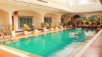 Una piscina con sillas de descanso y sombrillas, rodeada de plantas en macetas y un edificio con ventanas arqueadas.