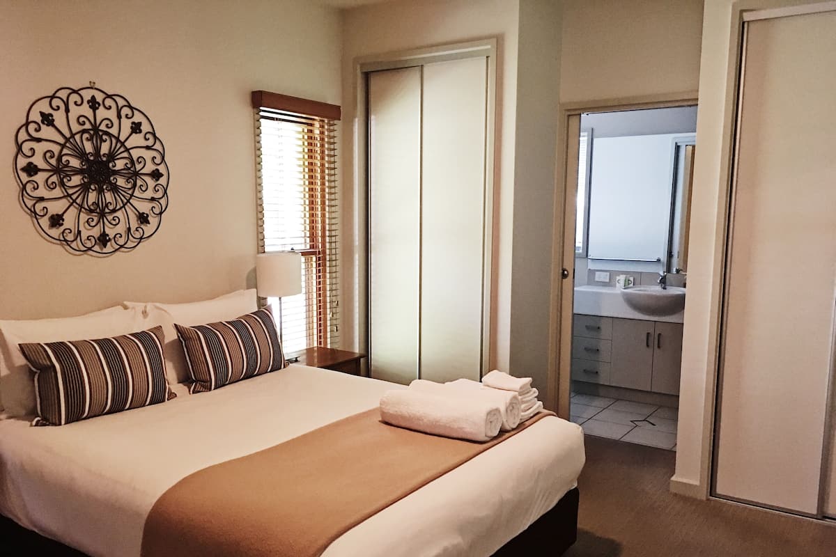 A neatly made bed with a brown throw, a decorative wall piece, and a bathroom visible through an open door.