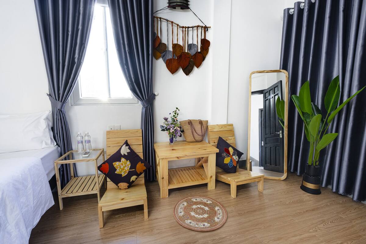 A room with a bed, two wooden chairs, a small table, a mirror, a hanging plant, and a window with curtains.