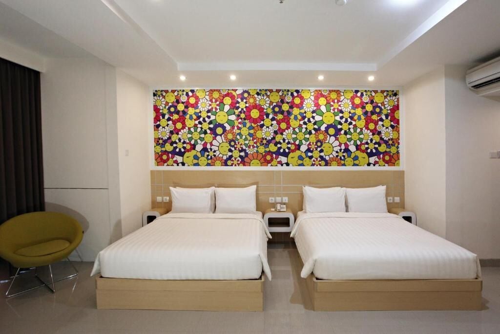 A hotel room with two beds, a colorful floral wall mural, a modern yellow chair, and a ceiling with recessed lighting.