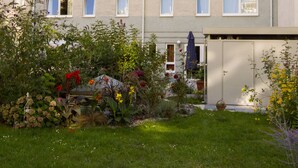 Un edificio a più piani con tetto piatto, diverse finestre e un piccolo giardino con varie piante e fiori.