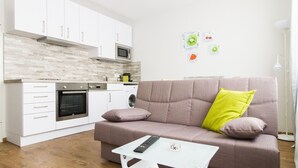 Una cucina moderna con mobili bianchi, un forno a microonde e una lavastoviglie. C'è un divano beige con un cuscino giallo brillante e un telecomando su un tavolino.