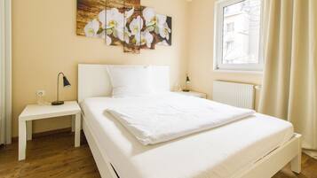 A bedroom with a large bed, white nightstands, a wall-mounted lamp, and a window with curtains.
