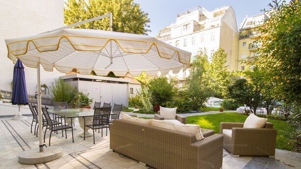 Patio esterno con mobili in vimini, un tavolo in vetro e un ombrellone bianco.