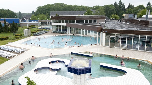 Zona de piscina con varias piscinas y un jacuzzi, rodeada de tumbonas y un edificio con grandes ventanales.