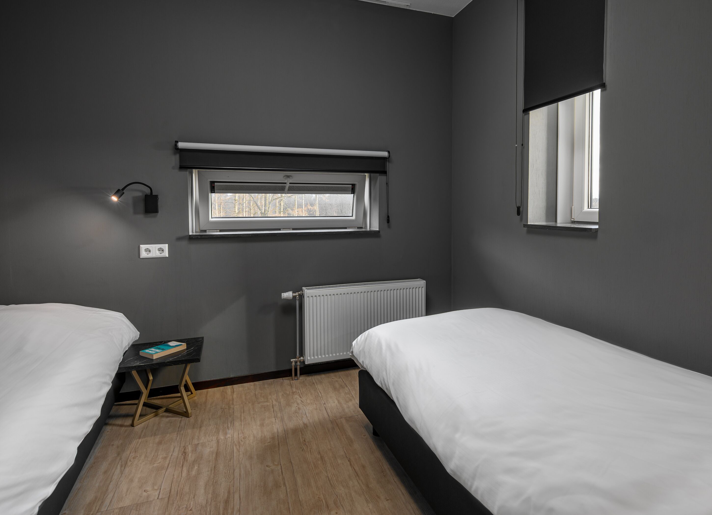 A bedroom with two beds, a window, a radiator, and a bedside table with a book.