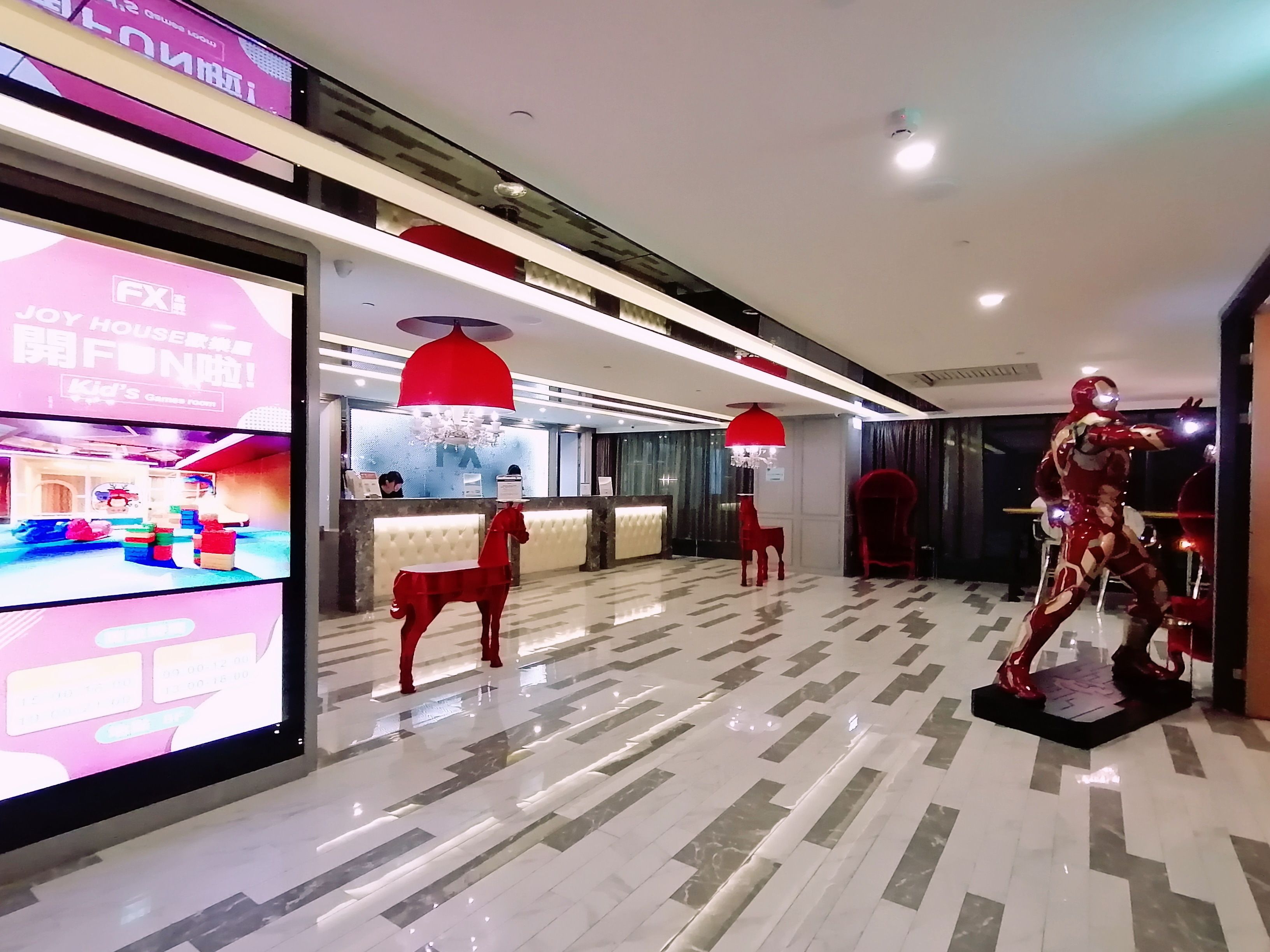 Un hall d’hôtel comprenant un comptoir d’accueil, une sculpture équestre rouge et un grand écran numérique affichant une salle de jeux pour enfants.