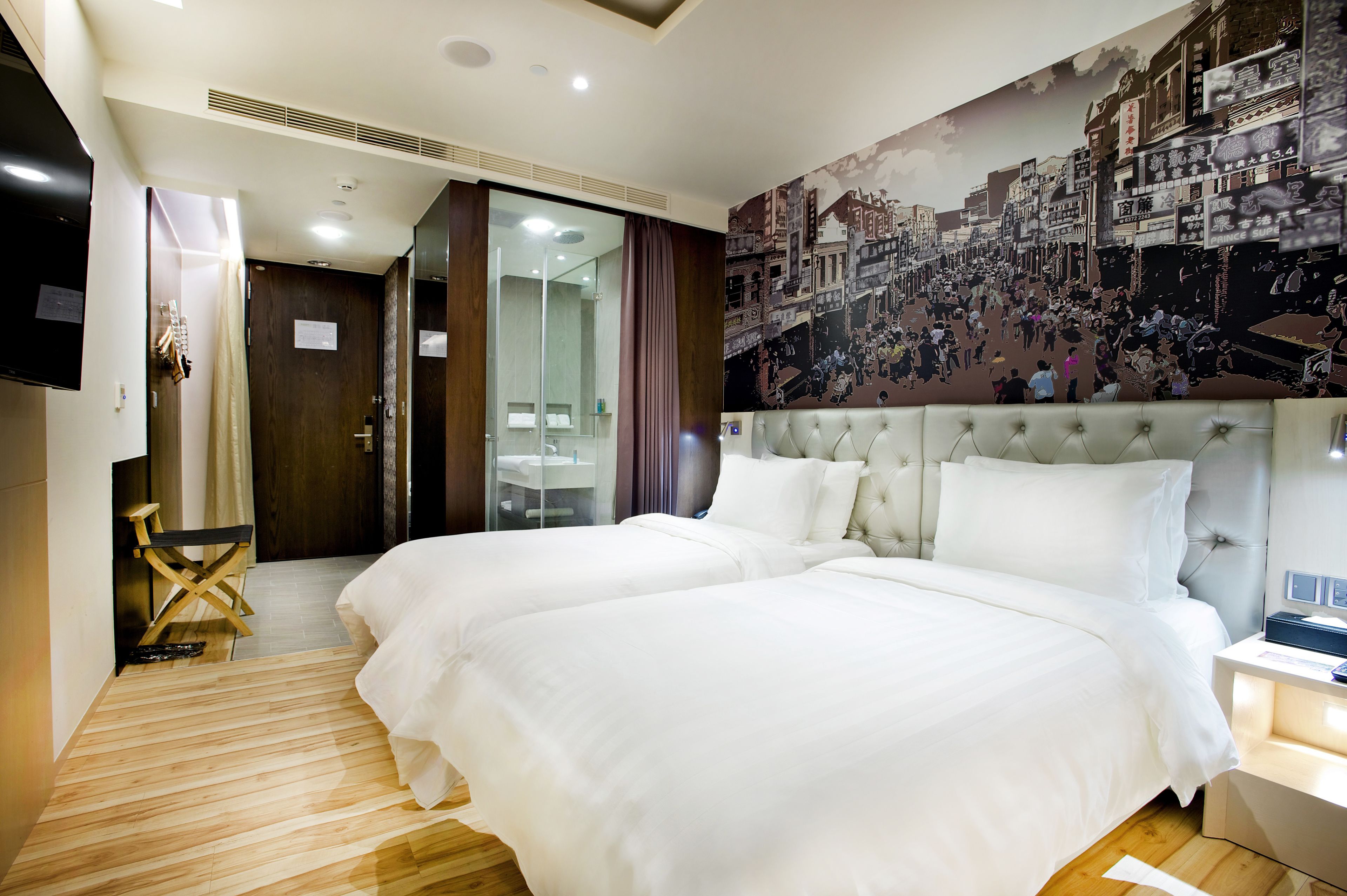 Habitación de hotel con dos camas, un mural de gran formato con un paisaje urbano, suelo de madera y un baño moderno con separador de vidrio.