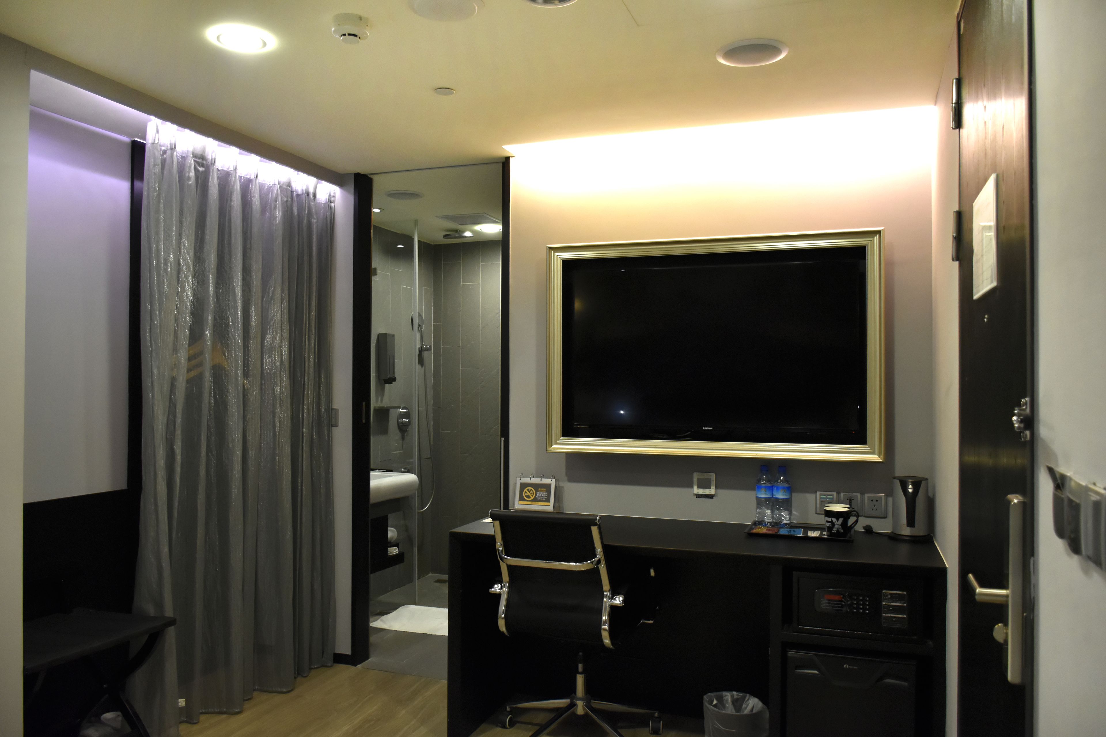 A hotel room with a desk, chair, and a large flat-screen TV. There is a shower area visible in the background.
