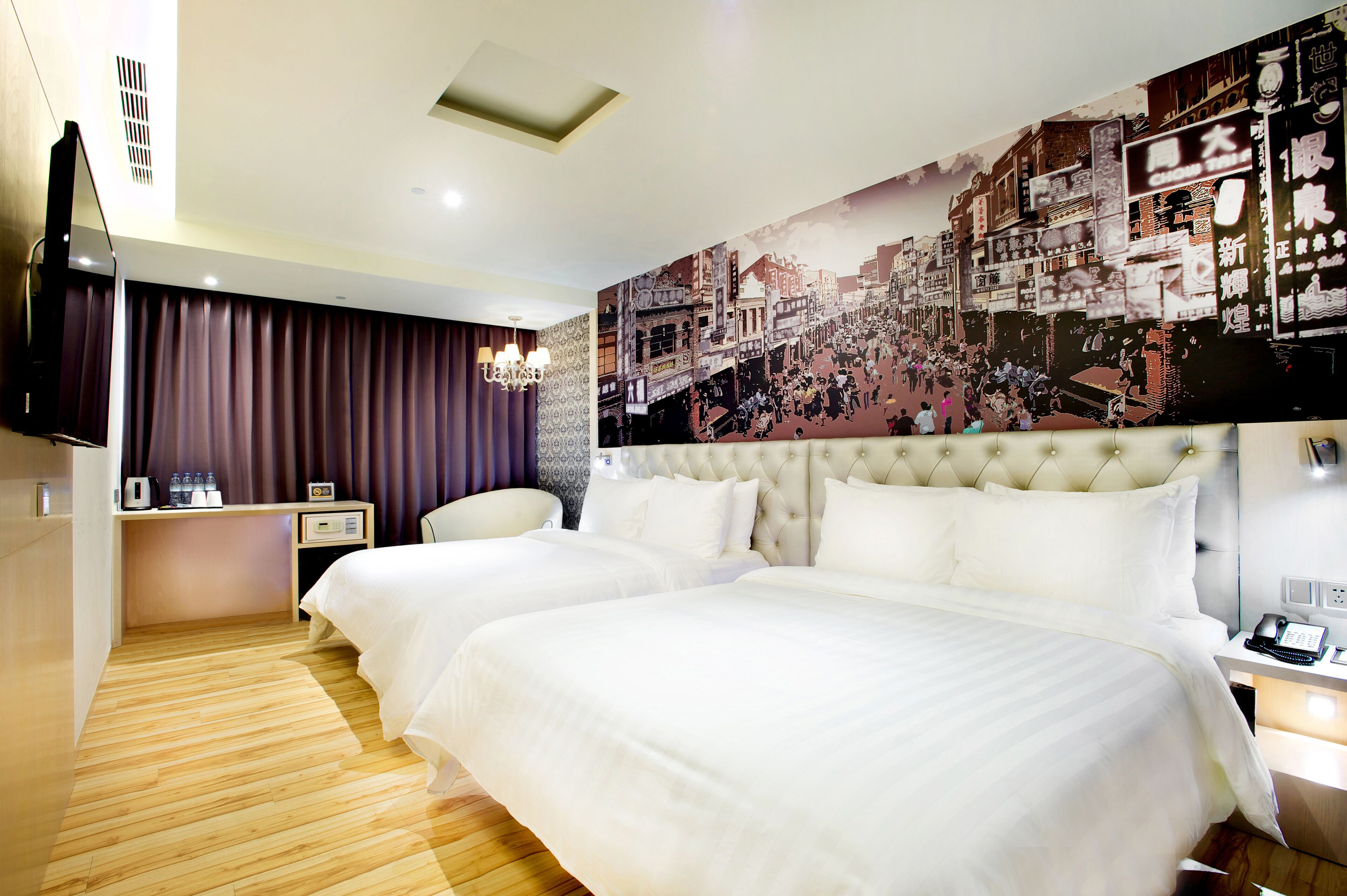 A hotel room with two beds, a large cityscape mural, a flat-screen TV, a bedside table with a lamp, and a wooden floor.