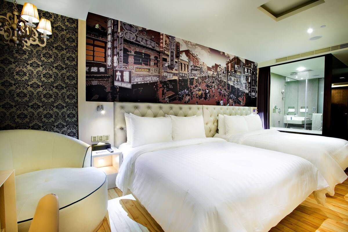 Hotellihuone, jossa on kaksi sänkyä, suuri kaupunkimaisemaa esittävä seinämaalaus, napitettu pääty ja puulattia.